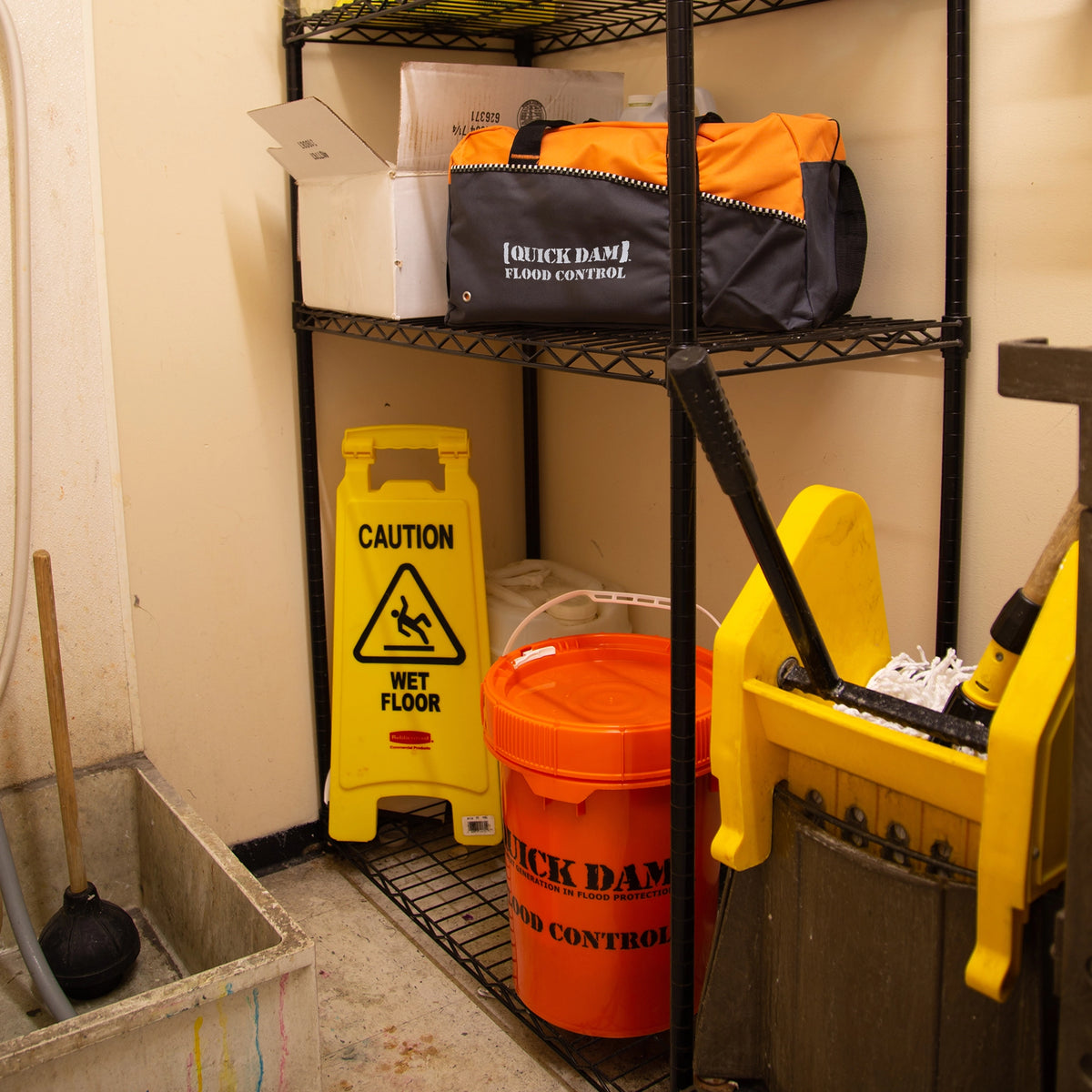 Quick Dam Flood Control duffle bag and bucket stored on a utility shelf in a janitor’s closet, ready for emergency flood protection.