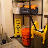 Quick Dam Flood Control duffle bag and bucket stored on a utility shelf in a janitor’s closet, ready for emergency flood protection.