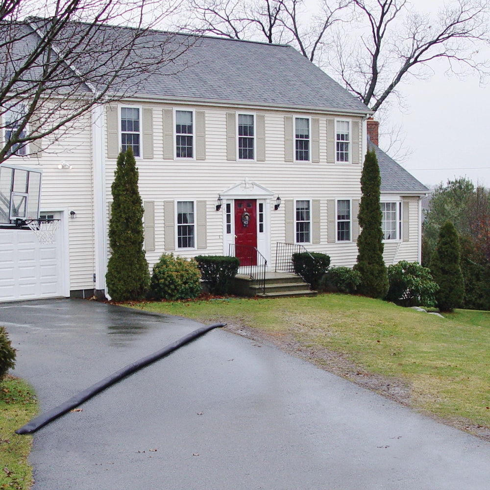Quick Dam Flood Barrier placed across a residential driveway to divert rainwater away from a two-story white house with red front door.