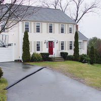 Quick Dam Flood Barrier placed across a residential driveway to divert rainwater away from a two-story white house with red front door.