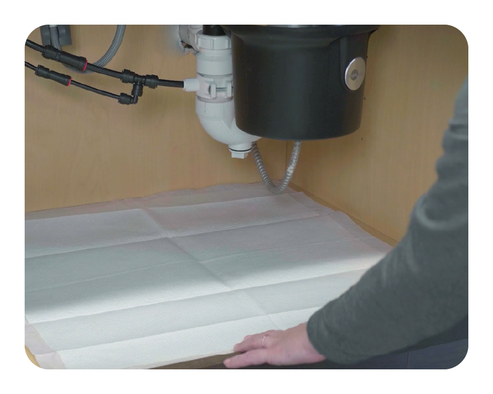 Person adjusting a drip mat under a kitchen sink with plumbing visible