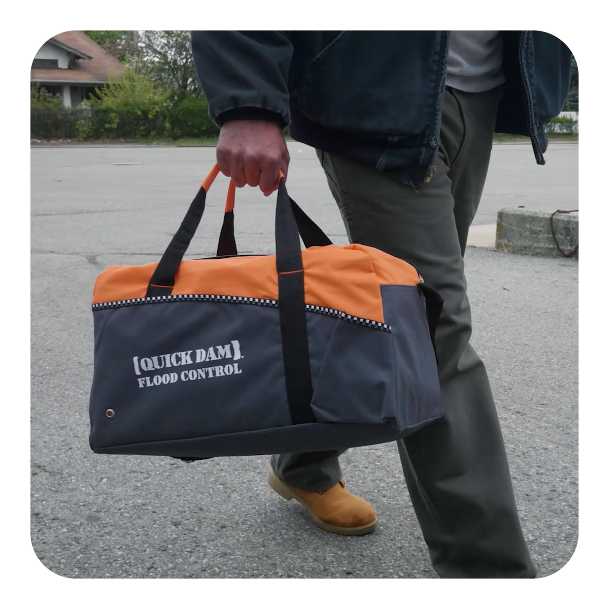 Person holding a black and orange bag with 'Quick Dam Flood Control' text on a street.