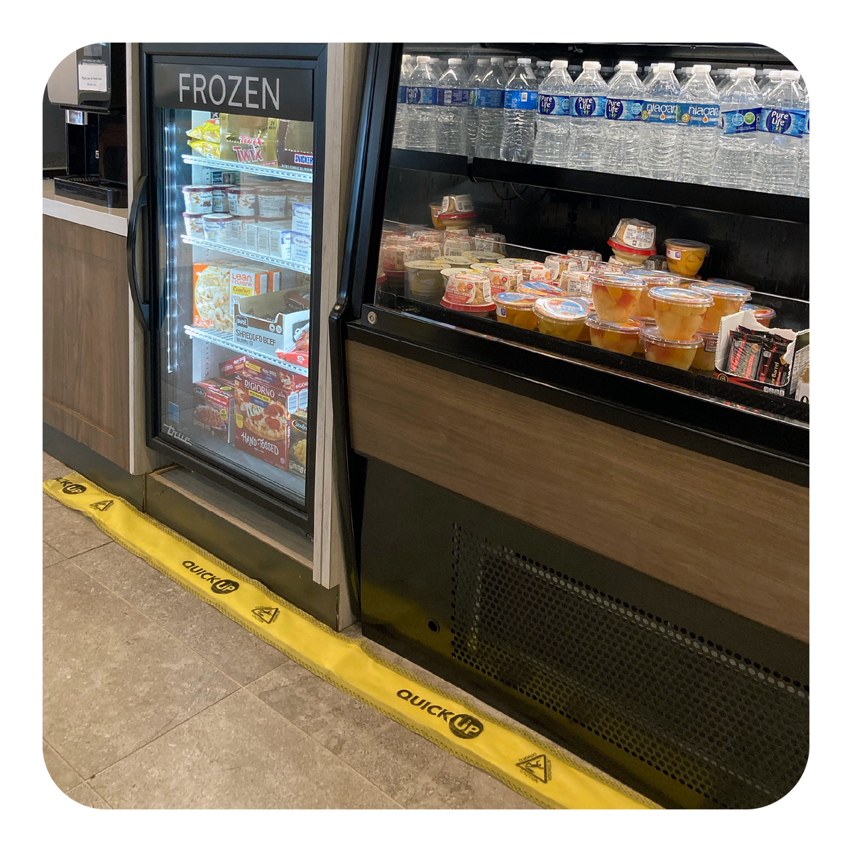 Frozen food and drink display case with water bottles and snacks in a store setting with a yellow Quick Up on the floor