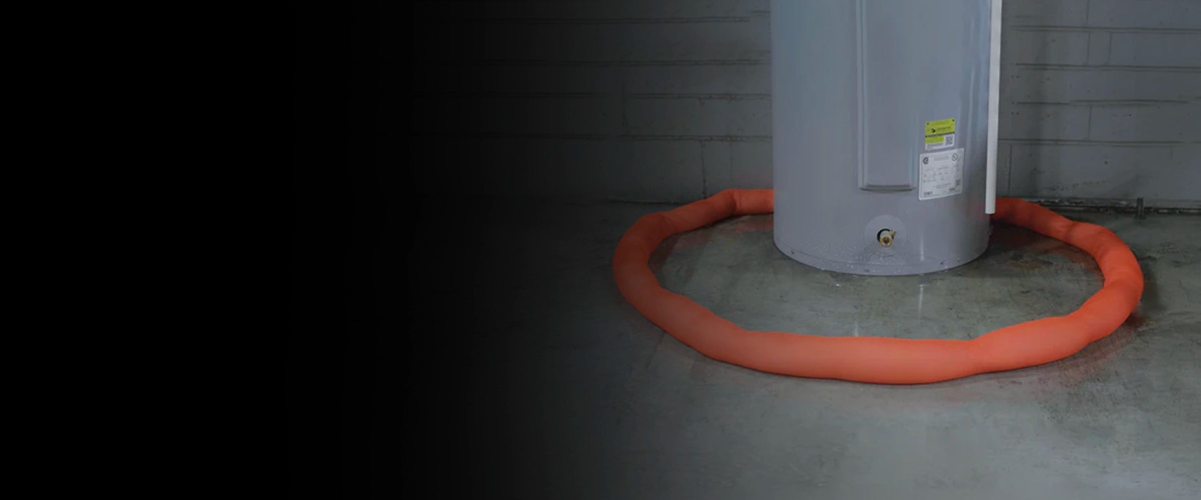 Gray water heater with an orange water dam on a concrete floor against a dark wall.